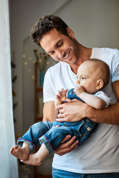 Handsome father with cute son in domestic room. Smiling father is carrying baby boy at home. They are in casuals.